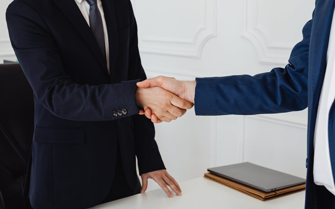 Two people in business attire shaking hands over a desk, after a deal acquiring private company